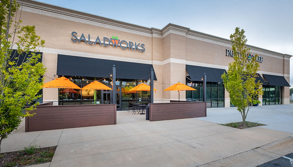 A Saladworks restaurant with outdoor seating under orange umbrellas, located in a modern shopping center with large black awnings and young trees nearby.