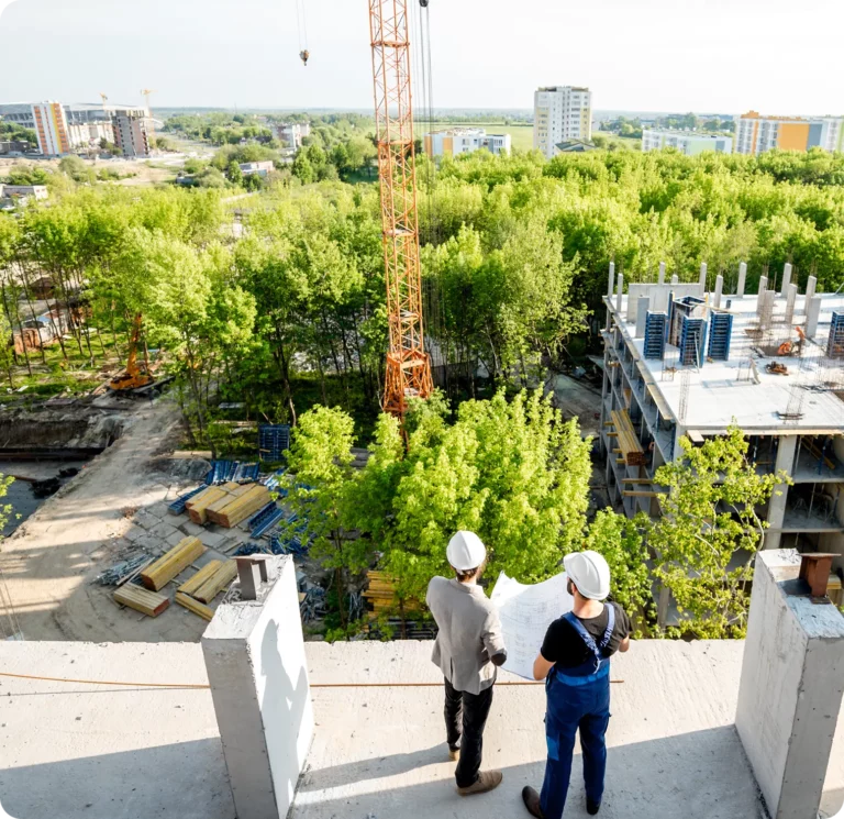 Two construction workers wearing safety helmets examine blueprints while overlooking a building site surrounded by trees and city buildings in this Placeholder Post.
