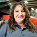 A woman with wavy brown hair smiles while wearing a dark gray polo shirt, standing indoors in an industrial setting.