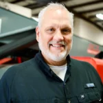 A middle-aged man with gray hair and a beard, wearing a black button-up shirt, smiles while standing indoors in an industrial setting.