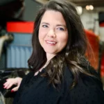 A woman with long brown hair and a black top smiles at the camera in an industrial indoor setting.