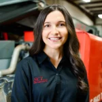 A woman wearing a black shirt with SCI embroidered on it stands indoors near industrial machinery, smiling at the camera.