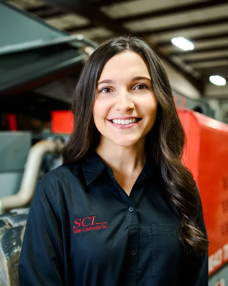 A woman with long brown hair wearing a black shirt with an SCI State Contractors Inc. logo stands indoors in front of industrial equipment.