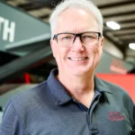 Smiling man with gray hair and glasses wears a dark polo shirt with an embroidered logo, standing indoors near industrial equipment.