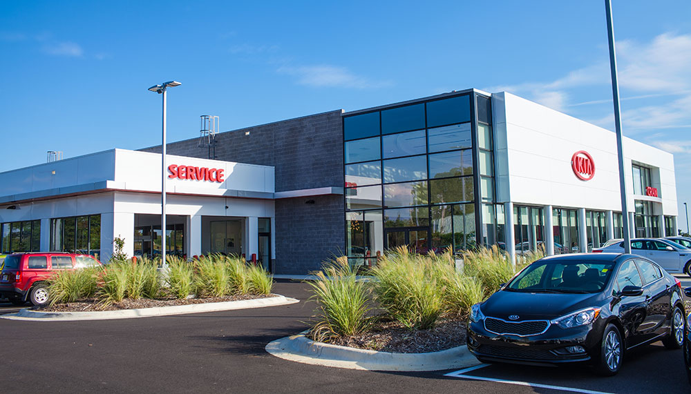 Modern Kia car dealership building with large windows, a Service sign, and several cars parked in the lot on a sunny day.