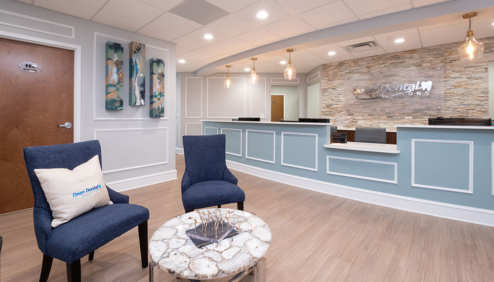 Modern dental office reception area at Dean Dental Solutions with blue chairs, a marble coffee table, wall art, and a front desk featuring pendant lights and a stone accent wall.