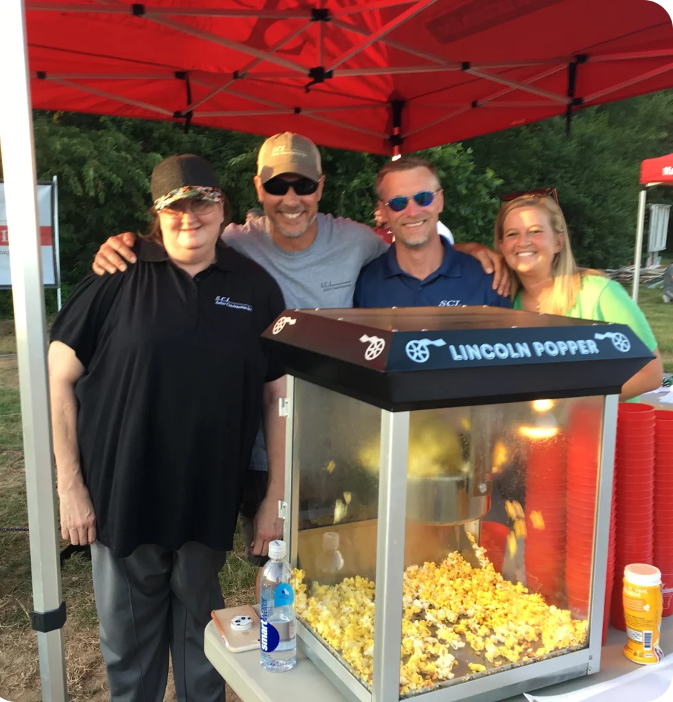 Four adults stand smiling behind a popcorn machine under a red canopy at an outdoor event, with drinks and supplies on the table in front of them.