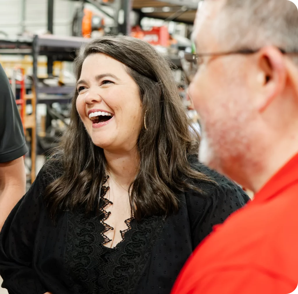 A woman with long brown hair and a black top laughs while talking with others in what appears to be a workshop or warehouse setting.
