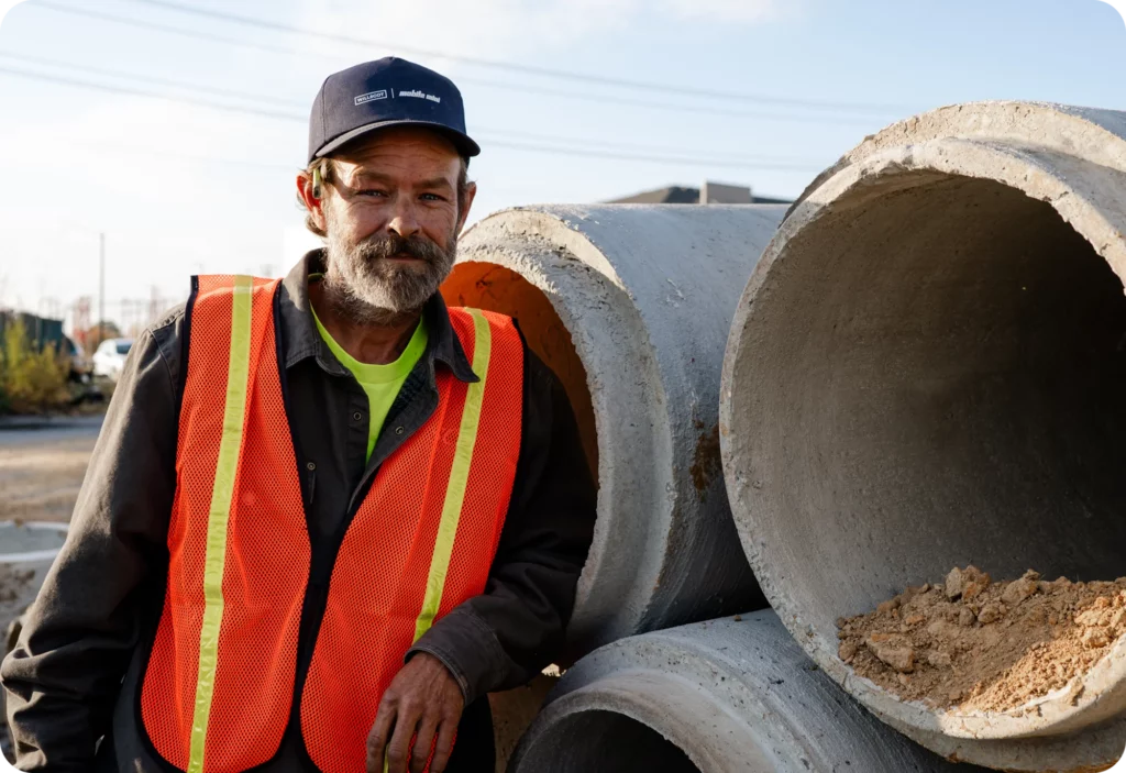 A construction worker in a safety vest and cap stands next to large concrete pipes at a worksite, with dirt inside one of the pipes.