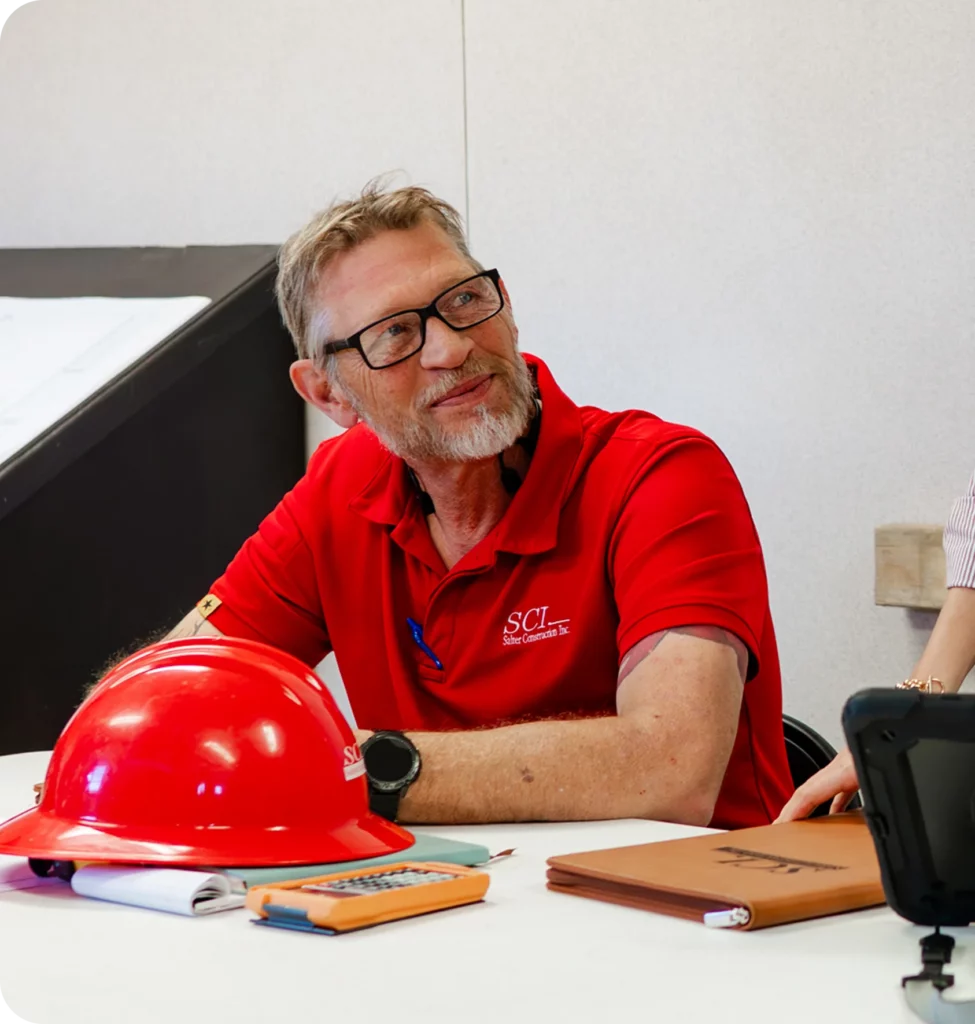 A man in a red shirt and glasses sits at a table with a red hard hat, notebook, and calculator in front of him, looking to the side and smiling.