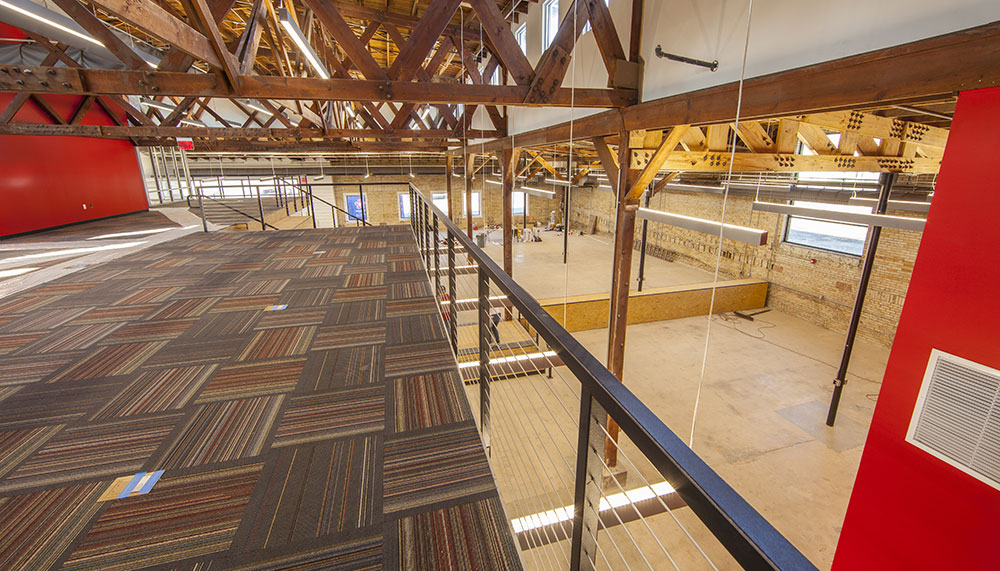 Interior view of a modern loft-style office space in Arkansas’s Regional Innovation Hub, featuring exposed wooden beams, a carpeted mezzanine, red accent walls, and large windows that fill this former Silver Mine with natural light.
