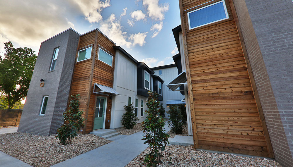 Modern T1 Townhomes with a mix of wood and brick exteriors, large windows, and landscaped with small trees and rocks, stand under a partly cloudy sky.