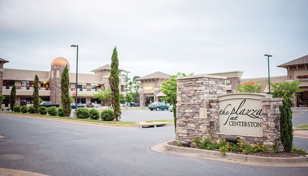 Shopping center parking lot with stone sign reading The Plaza at Centerstone, surrounded by stores, trees, and lamp posts on a cloudy day at Centerstone.